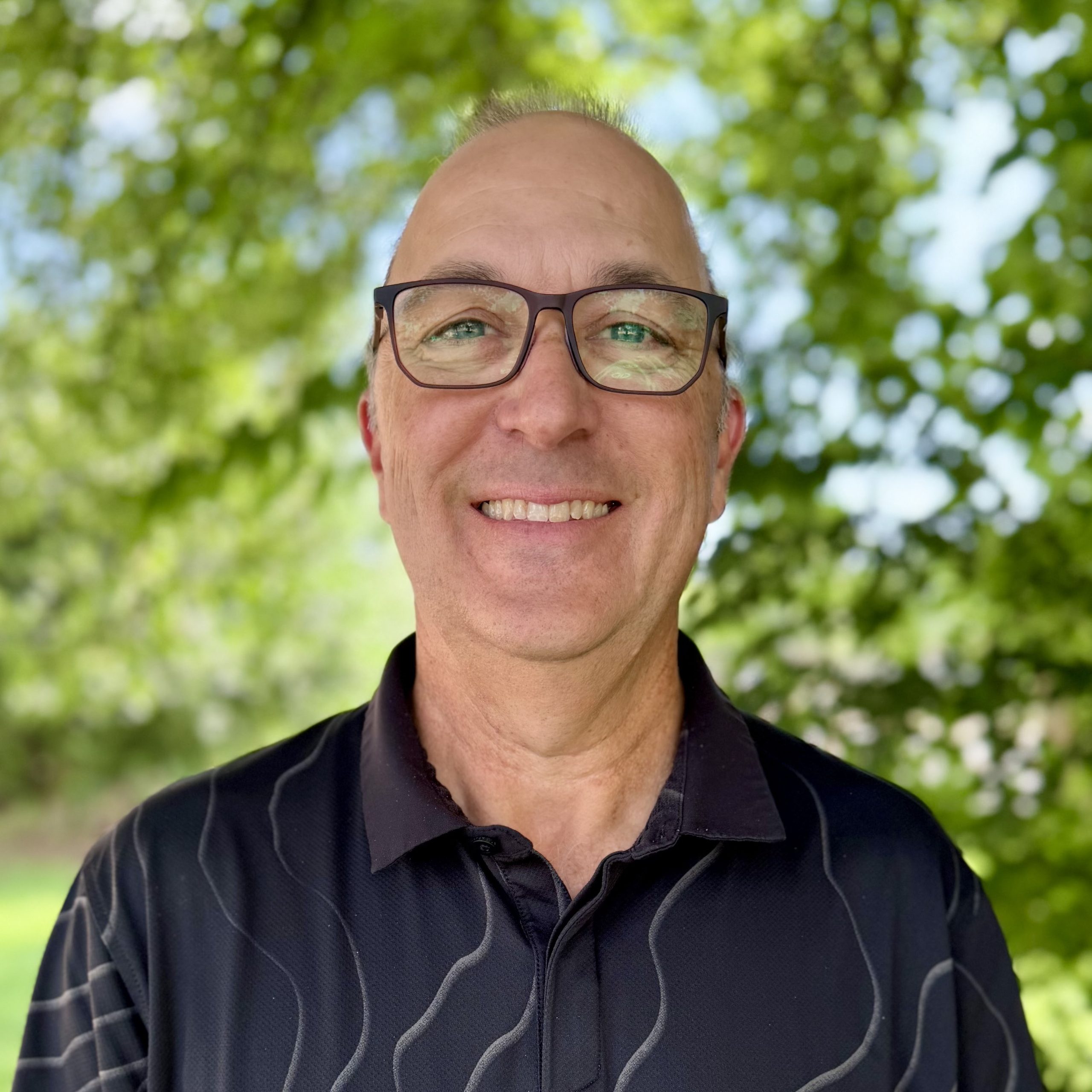 A smiling man in dark-rimmed glasses and wearing a dark gray shirt with a wavy, light gray pattern