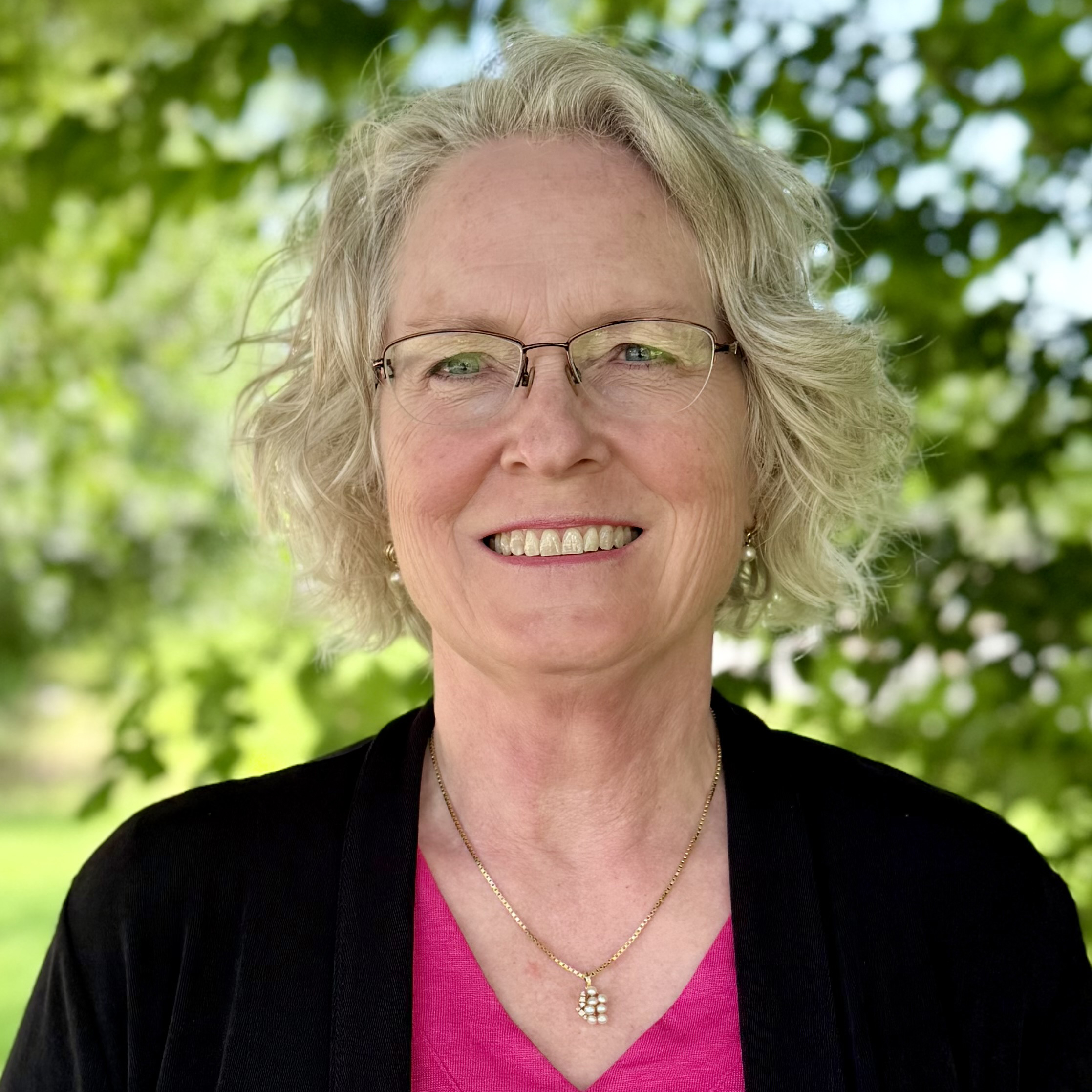 A smiling woman in glasses, wearing a bright pink shirt and a black cardigan