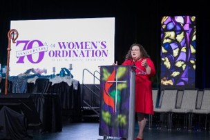 Woman speaking infront of sign reading, 70th Anniversary of Women's Ordination