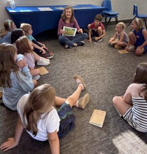 A woman and a group of children sit in a circle on the floor.