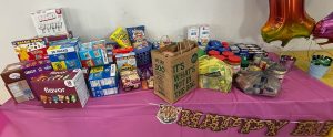 An assortment of donated nonperishable food items sit on a table covered with a pink tablecloth.